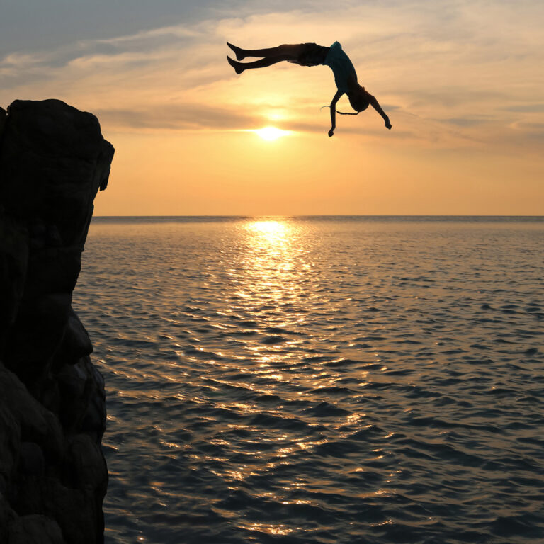 Person diving, doing a gainer off a tall cliff at sunset in a golden sky