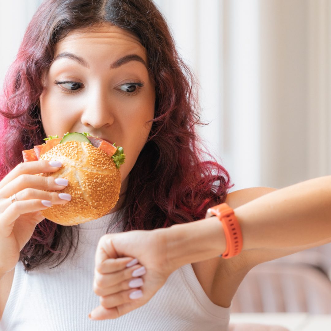 Young woman eating a sandwich and looking to the side at her watch